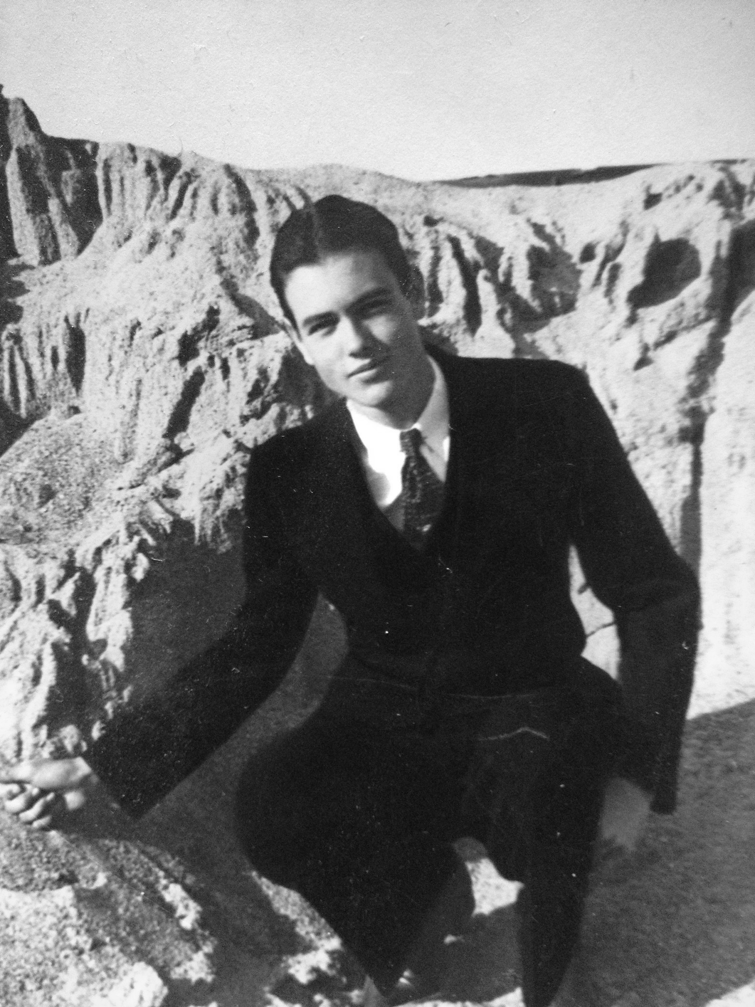 A black and white photo of a young Milton Greer, crouching in a suit against a sandy slope